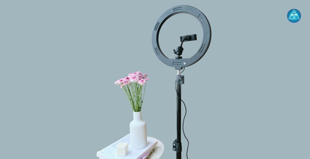 Ein schwarzes Ringlicht auf einem Stativ mit Smartphone-Halterung steht rechts neben einem kleinen Tisch, auf dem Bücher, eine weiße Kerze und eine weiße geriffelte Vase mit rosa Blumen liegen, vor einem graublauen Hintergrund mit dem runden blauen Logo von Dein Gesundheitsbusiness oben rechts.