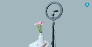 Ein schwarzes Ringlicht auf einem Stativ mit Smartphone-Halterung steht rechts neben einem kleinen Tisch, auf dem Bücher, eine weiße Kerze und eine weiße geriffelte Vase mit rosa Blumen liegen, vor einem graublauen Hintergrund mit dem runden blauen Logo von Dein Gesundheitsbusiness oben rechts.