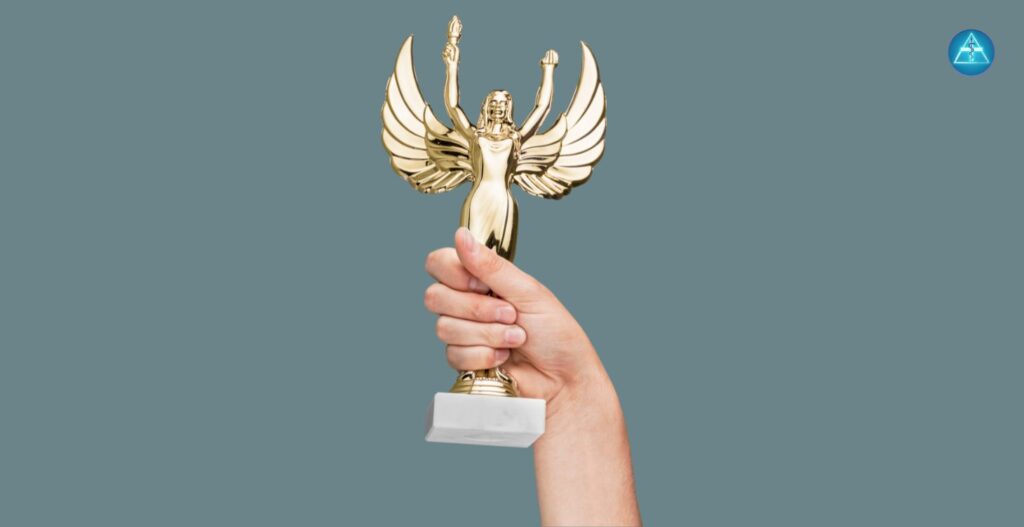 Ein nach oben gestreckter Arm hält eine goldene Trophäe mit Engelsflügeln vor einem graublauen Hintergrund, in der oberen rechten Ecke ist das Logo von Dein Gesundheitsbusiness zu sehen.