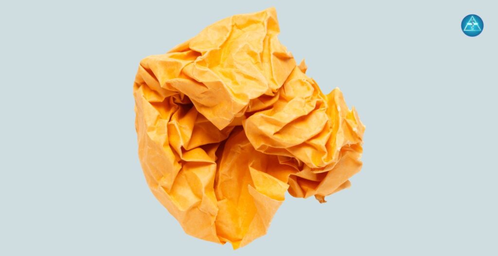 Alt-Text: Eine zerknüllte gelborange Papierkugel vor einem hellblauen Hintergrund mit dem runden blauen Logo von Dein Gesundheitsbusiness in der oberen rechten Ecke.
