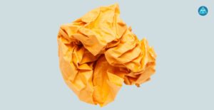 Alt-Text: Eine zerknüllte gelborange Papierkugel vor einem hellblauen Hintergrund mit dem runden blauen Logo von Dein Gesundheitsbusiness in der oberen rechten Ecke.