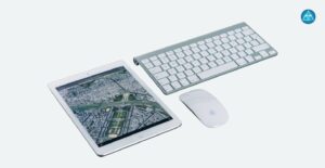 Ein weißes Tablet mit einer geöffneten Satelliten-Stadtkarte auf dem Bildschirm liegt schräg neben einer kabellosen silber-weißen Tastatur und einer weißen Computermaus vor einem hellgrauen Hintergrund, während oben rechts das runde blaue Logo von Dein Gesundheitsbusiness zu sehen ist.